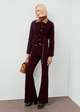 Pantalón jeans flare de terciopelo color bordó, de tiro alto y diseño largo, con cinco bolsillos, trabillas para cinturón y cierre frontal con botón y cremallera.