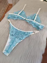 Conjunto de lencería compuesto por un corpiño triangular y una bombacha, confeccionados en encaje floral color aguamarina con detalles de ribetes en color champagne. El corpiño cuenta con breteles regulables y la bombacha presenta laterales ajustables.