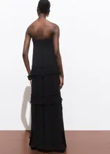 Vestido maxi largo negro de viscosa, palabra de honor y sin mangas. Presenta diseño evasé con detalle de volantes escalonados y cierre de cremallera lateral.