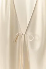 Camisa de tejido satinado con escote en pico y manga larga. Presenta un diseño holgado con cierre frontal mediante lazada y detalle de aplicación de encaje a tono en el ruedo.