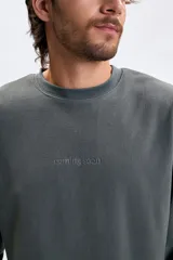 Buzo de polar gris oscuro con cuello redondo, mangas largas y puños elásticos. Presenta un estampado minimalista con la frase 'coming soon' en el centro del pecho.