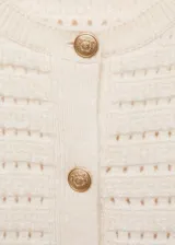 Chaqueta de punto calado en color crema, con cuello redondo, manga larga y cierre frontal mediante botones dorados. Presenta un diseño recto y tejido de punto medio.
