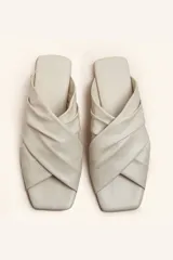 Sandalia tipo slide de cuero vacuno color blanco, con diseño de tiras cruzadas y fruncidas. Presenta una puntera cuadrada y una suela plana de cuero con un pequeño taco de 2 cm.