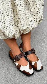 Zapatos tipo Mary Jane con estampado de vaca, confeccionados en cuero con textura de pelo. Presentan una tira sobre el empeine con hebilla metálica ajustable y punta cuadrada.