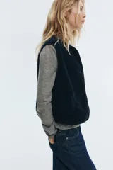 Chaleco de sherpa color negro, con cuello redondo y cierre frontal de botones a presión. Presenta bolsillos laterales de parche y ribetes finos en los bordes.