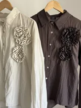 Camisa de manga larga con cuello clásico, confeccionada en tela nacarada con brillo sutil. Presenta apliques de flores tridimensionales fruncidas en el frente, una en color beige claro y dos en color marrón oscuro.