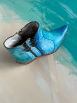 Bota corta de cuero vacuno pintada a mano en tonalidades turquesa y celeste, con taco cuadrado de madera, doble cierre lateral con tiradores metálicos personalizados y punta fina.