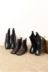 Botas tipo chelsea de cuero color marrón oscuro con acabado brillante. Presentan punta cuadrada con detalle de puntera metálica, elásticos laterales y taco cuadrado bajo.