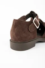 Sandalia tipo cangrejera de gamuza color marrón, con diseño de tiras entrelazadas, cierre con hebilla metálica lateral y suela plana de goma.