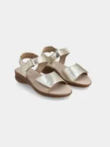 Sandalia de cuero con tiras ajustables de velcro en el empeine y el talón. Presenta un diseño bicolor en tonos dorado y beige, con una suela de goma texturizada de aspecto natural.
