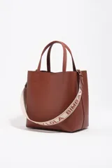 Bolso de hombro de tamaño mediano color marrón, confeccionado en piel vacuna. Presenta doble asa corta de mano y correa de tejido jaspeada con el nombre de la marca estampado. Incluye detalles en piel, cierre con botón de presión, logo grabado en dorado en el frente y acabados metálicos.