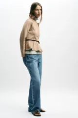 Chaqueta color beige con efecto ante, cuello redondo, manga larga y cierre frontal con cremallera. Presenta un bajo en evasé y un cinturón fino de cuero marrón oscuro con hebilla metálica.