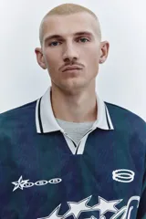 Camiseta de fútbol de manga corta en malla estampada, con diseño a rayas verticales en tonos azul y verde oscuro. Cuenta con cuello polo blanco y escote en V con ribete negro, además de estampados gráficos frontales con estrellas y la inscripción "Run Wild". Presenta un corte holgado.
