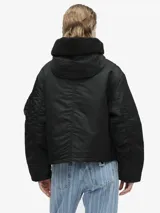 Chaqueta corta tipo bomber con capucha, color verde militar, con cierre frontal de cremallera y botones, bolsillos laterales con solapa y bolsillo utilitario en la manga.