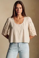 Blusa color arena de algodón peinado, con escote redondo, mangas cortas abullonadas con bordados suizos y detalles de alforcas en el frente.