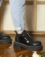Botinetas negras de caña baja con suela track dentada. Presentan un diseño combinado en cuero texturizado y paneles de charol brillante, con cierre frontal metálico y costuras visibles en el borde de la suela.
