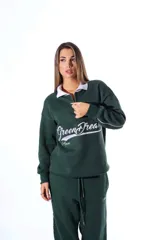 Polo de frisa color verde oscuro con cuello blanco y cierre parcial frontal. Presenta un bordado en el frente con el texto "Green Dreams" en tipografía cursiva blanca y la palabra "Aum" debajo.