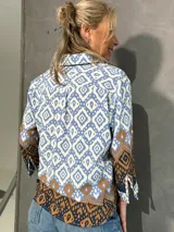 Blusa de manga larga con cuello camisero, corte recto y cierre frontal de botones. Presenta un diseño estampado geométrico en tonos celeste, blanco y marrón.