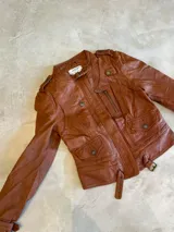 Chaqueta biker corta confeccionada en 100% cuero genuino ovino, color camel. Cuenta con cuello mao con tira y hebilla metálica, bolsillos frontales con cierre, detalles de trabillas con botones y tiras ajustables en los puños.