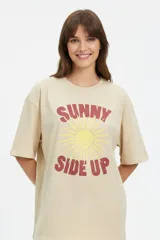 Remera de algodón color beige, de manga corta, con cuello redondo y diseño oversize. Cuenta con un estampado frontal que dice "SUNNY SIDE UP" en tonos rojo y amarillo, acompañado de un gráfico de sol sonriente.