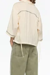 Camisa corta de corte boxy en color marrón, con cuello camisero, cierre frontal de botones y mangas 3/4. Presenta pespuntes decorativos a contraste en los puños y una textura sutil en el tejido.