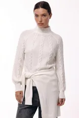 Polera de tejido de punto color blanco con diseño de trenzas verticales, cuello alto y mangas largas con puños ajustados. Presenta un detalle de lazo lateral integrado en la cintura.