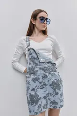 Jumper de jean corto, de color celeste claro con estampado floral en tonos oscuros. Presenta tirantes regulables con hebillas metálicas, bolsillo pectoral y bolsillos laterales funcionales.