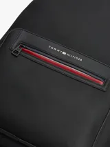 Mochila Tommy Hilfiger de color negro, confeccionada en poliuretano con paneles de texturas mixtas y diseño curvo. Cuenta con un compartimento principal con cremallera de doble cierre, funda interior para portátil, bolsillo interior con cremallera y un bolsillo exterior frontal con cierre y detalles en rojo y azul marino. Incorpora asa superior, correas ajustables y acolchadas, respaldo con panel de malla acolchado y logo metálico de la marca en la parte delantera.