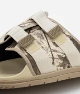 Ojota tipo slide con estampa camuflada en tonos beige y marrón. Presenta dos tiras ajustables con cierre de velcro y suela de goma antideslizante.