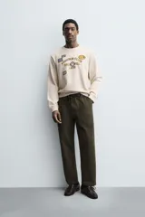 Sudadera de cuello redondo y manga larga en color beige. Presenta un diseño con texto bordado y múltiples parches decorativos en el frente, con terminaciones en rib en puños y cintura.