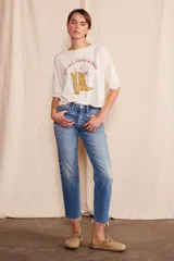 Remera de algodón orgánico color blanco hueso, con cuello redondo y mangas cortas dobladas. Presenta un estampado frontal con botas de cowboy amarillas y un sombrero rosa, y el texto "Not My First Rodeo".