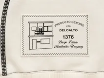 Buzo blanco de manga larga con diseño holgado, cuello redondo y costuras externas visibles en contraste. Cuenta con una etiqueta decorativa en el frente inferior.