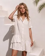Vestido camisero corto de lino color blanco, con mangas cortas, cierre frontal de botones, cuello camisero y volados asimétricos en el ruedo.