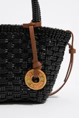 Bolso tipo cesta de estructura rígida y diseño trenzado en color negro. Cuenta con dos asas superiores cortas y una correa larga ajustable en color marrón. Incluye un dije circular decorativo con grabado en tono madera.