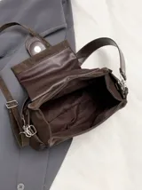 Cartera de hombro color marrón oscuro, con diseño estructurado y detalle de hebilla central decorativa con ojales metálicos. Incluye correa de hombro fija y correa larga ajustable y extraíble.