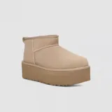 Botas de caña baja tipo botineta con plataforma elevada, confeccionadas en gamuza color marrón. Presentan costuras con detalle de puntada overlock, lengüeta trasera y forro interior de lana suave.