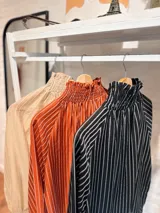 Blusa de tela liviana con diseño de rayas verticales en tonos beige y crema. Presenta cuello alto con detalle de fruncido tipo smock, mangas largas abullonadas con puños elásticos y volados en los extremos.