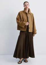 Chaqueta corta de algodón color marrón tostado, con cuello alto, cierre frontal de botones, bolsillos ribeteados y mangas largas con trabillas ajustables en los puños.
