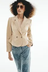 Blazer corto de cuero en color tostado, con cuello solapa, hombreras y manga larga. Presenta un diseño evasé en el bajo y un cierre frontal cruzado con dos botones.