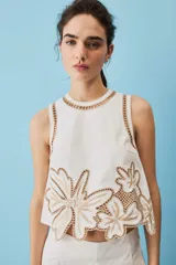 Blusa sin mangas de corte crop en color blanco, con detalles de bordados calados en tono tostado formando diseños florales en el cuello, los hombros y el borde inferior.