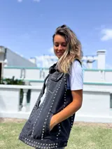 Chaleco de jean color negro gastado, con corte clásico y detalle de tachas metálicas plateadas distribuidas a lo largo de las costuras frontales y el ruedo.