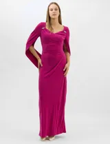 Vestido largo de seda fría color fucsia, con escote cruzado drapeado y apliques de pedrería en los hombros. Presenta mangas largas abiertas estilo capa y un drapeado lateral en la cintura que estiliza la silueta.