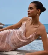 Vestido midi de lentejuelas color rosado, con escote drapeado en la parte delantera y escote degagê en la espalda con breteles finos cruzados. El bajo es asimétrico con punta y tiene un forro corto que crea un efecto de transparencia en las piernas.