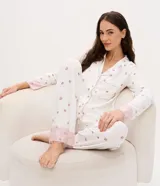 Pijama de dos piezas estilo americano, compuesto por camisa de manga larga con cuello en V, botones frontales y bolsillo en el pecho, y pantalón largo con cintura elástica. Presenta un estampado floral delicado sobre fondo blanco y detalles de encaje rosado en los puños y el ruedo del pantalón.