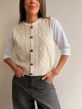 Chaleco tejido de punto color beige con diseño de rombos y trenzas en el frente. Presenta cuello redondo y cierre frontal con botones.