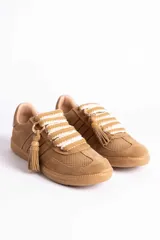 Zapatillas urbanas color tostado con textura geométrica en el cuerpo y detalles de gamuza en la puntera y laterales. Presentan un diseño distintivo con cordones anchos tejidos en tonos tostado y blanco, y un accesorio metálico con borla de gamuza en el empeine.