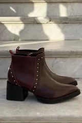Bota corta de cuero color bordó, con taco cuadrado negro y suela de plataforma. Presenta detalles de tachas metálicas plateadas a lo largo de la costura frontal y lateral, con cierre interno.
