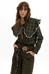 Camisa de manga larga confeccionada en popelina de algodón con estampado de cuadros en tonos verde oscuro y amarillo. Presenta un cuello amplio con bordes ondulados y terminaciones en encaje de algodón crudo, cierre frontal con botones de nácar y lazo de ajuste en el cuello.