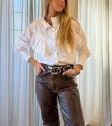 Camisa blanca de mangas largas con puños abotonados, cuello clásico y cierre frontal con botones. Presenta un volante con volados en diagonal desde el hombro hasta el centro.