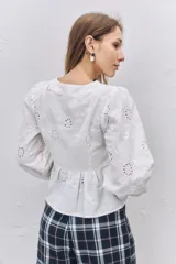 Blusa blanca de tela broderie con diseño de flores caladas, mangas largas abullonadas con puños elásticos y cierre frontal mediante lazos decorativos.